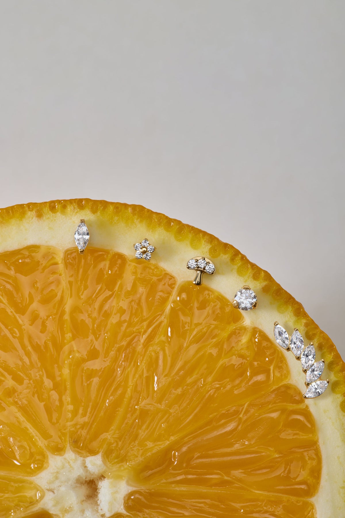 A close-up of a sliced orange displays the Marquise Threadless Flat Back Titanium Cartilage Stud in Gold, along with other sparkling gemstone earrings, arranged along the curved edge against a neutral background.