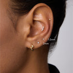 A close-up of a person’s ear with multiple piercings, including a 14K Solid White Gold Lab-grown Diamond Prong Set Flat Back Stud (1.5mm), plus gold and silver studs and a gold hoop. The person has medium brown skin and dark hair.