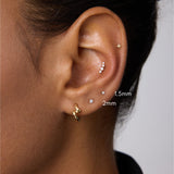 A close-up of a person’s ear with multiple piercings, including a 14K Solid White Gold Lab-grown Diamond Prong Set Flat Back Stud (1.5mm), plus gold and silver studs and a gold hoop. The person has medium brown skin and dark hair.