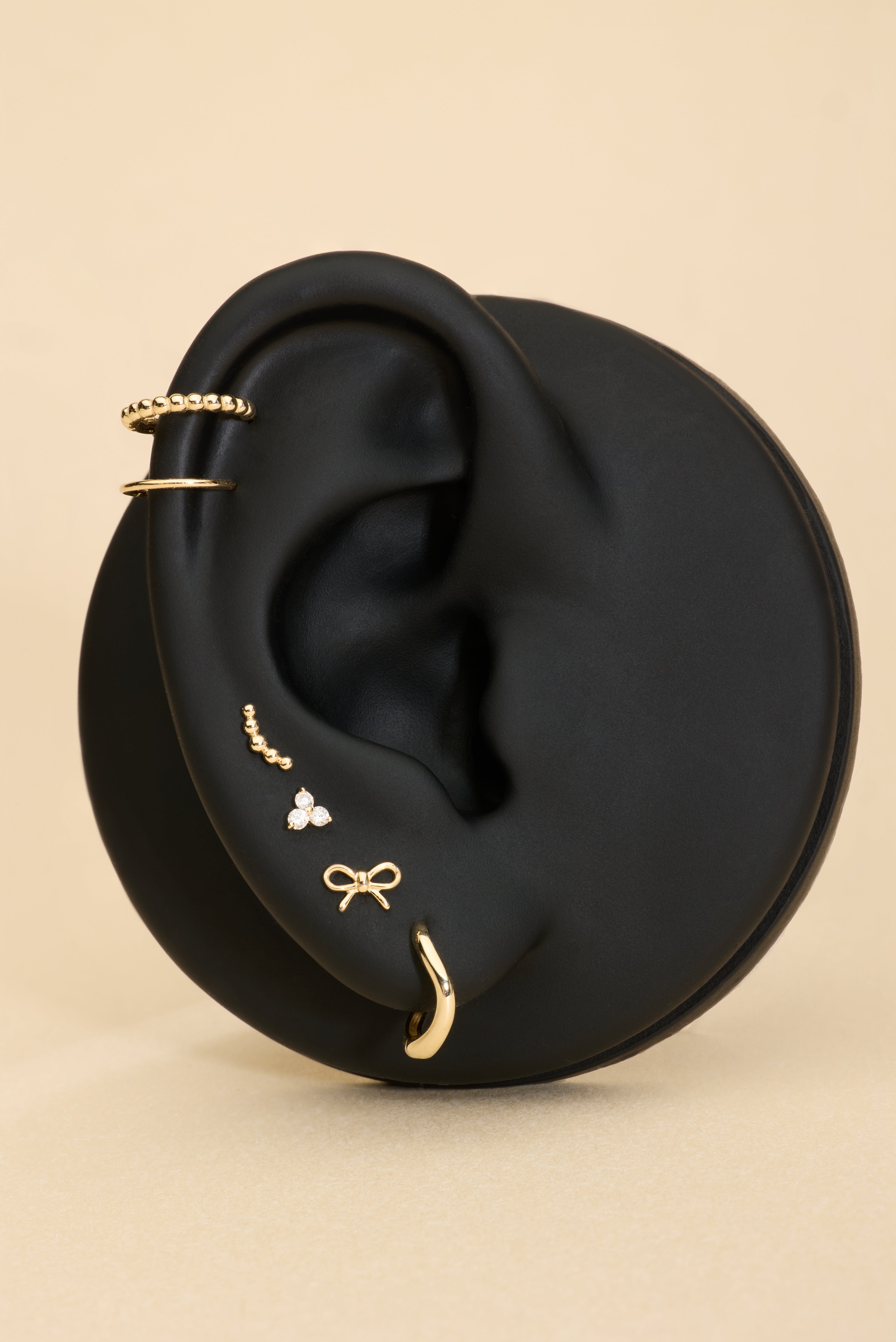 A black mannequin ear displays 14K Recycled Solid Yellow Gold Twisted Huggie Earrings among other gold styles against a beige background.