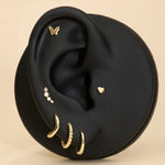 Black ear with gold earrings, butterfly flatback studs, heart flatback and diamond a beige background