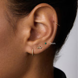 A close-up of an ear with four earrings: a 14k gold small hoop, a leaf-shaped stud, the 14K Solid Yellow Gold Green Emerald Flat Back Stud - 2mm, and a thin hoop on the upper ear. The person wears a black collared shirt.