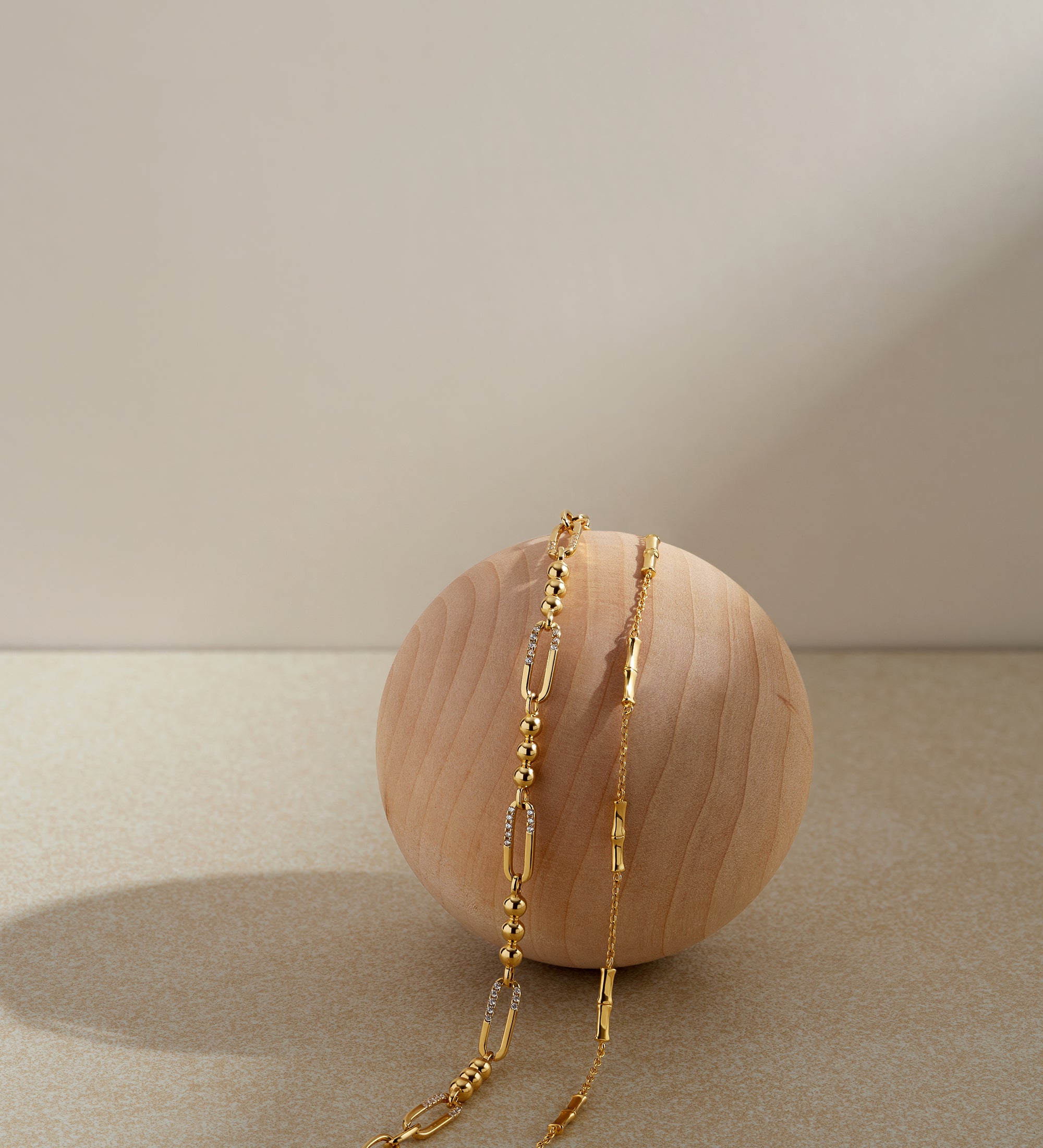 The Bamboo Chain Bracelet in Gold, featuring a delicate 18k gold-plated design, is elegantly displayed draped over a smooth, light wooden sphere against a neutral beige background with soft lighting and gentle shadows.