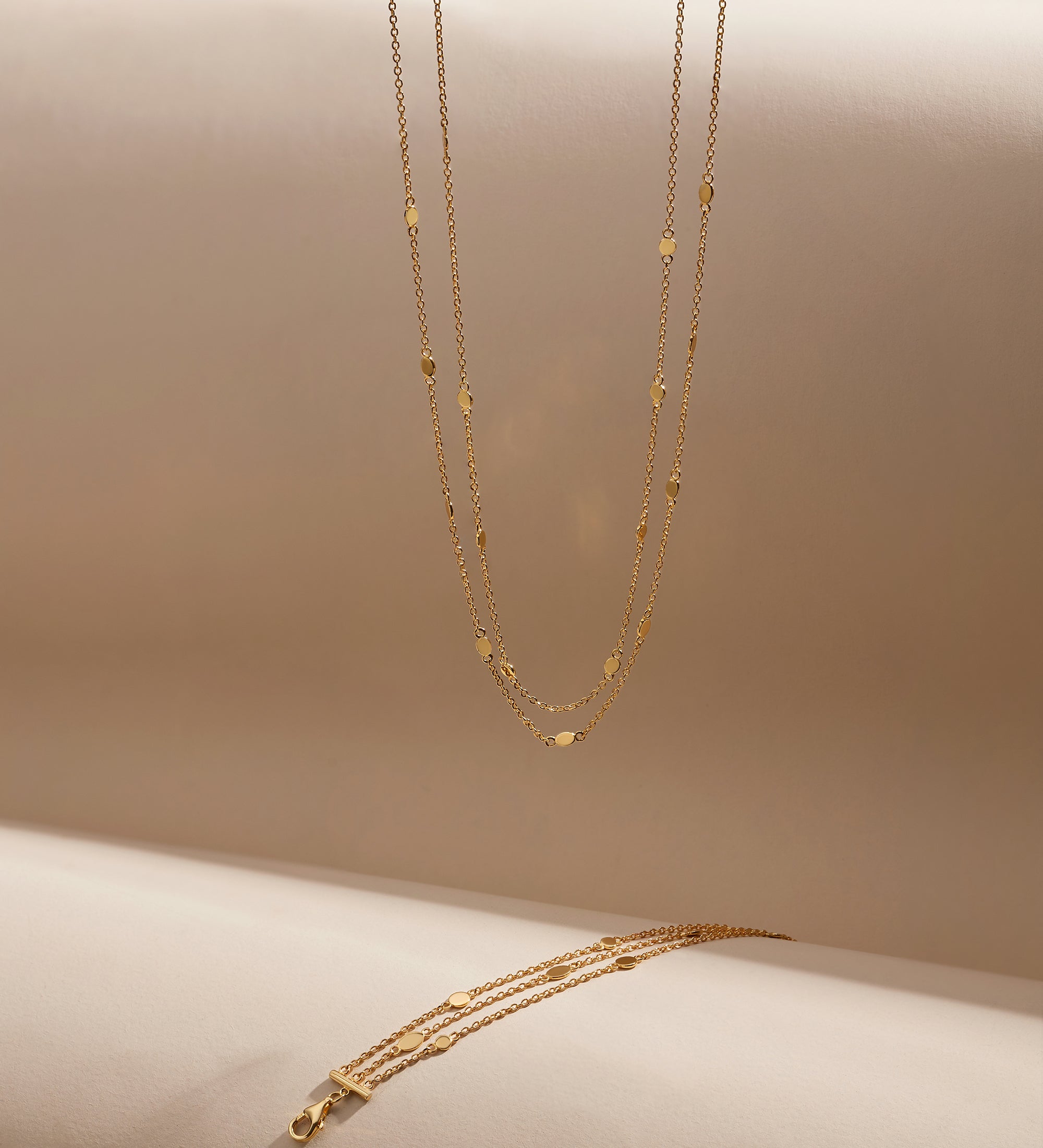 The Simona Double Chain Stacking Necklace in Gold features delicate double chains with small bead accents, displayed vertically and horizontally against a soft beige background.