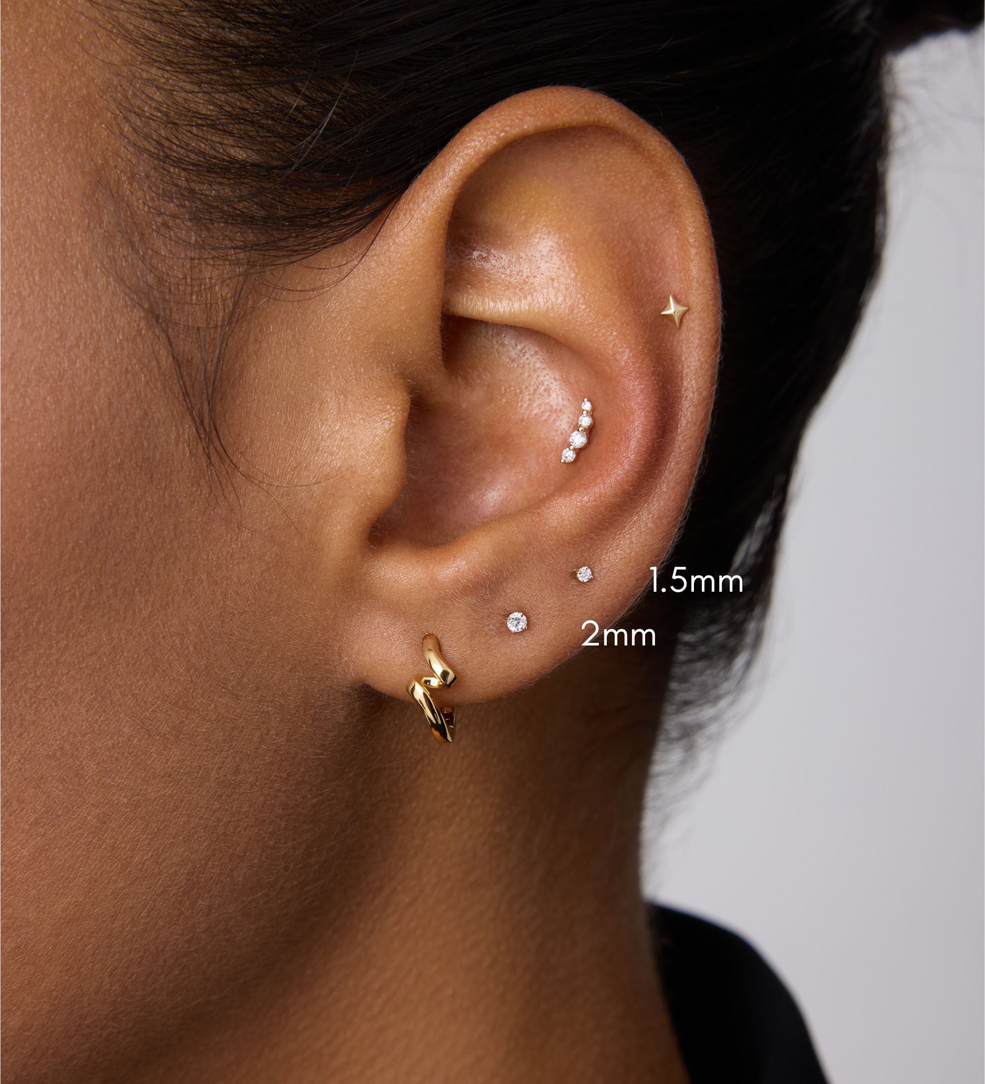 A close-up of a person’s ear with multiple piercings, featuring the 14K Solid Yellow Gold Lab-grown Diamond Prong Set Flat Back Stud - 1.5mm among other gold pieces. The person has medium brown skin and dark hair pulled back.