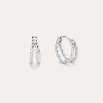 The Mae Twist Double Huggie Earrings in Silver feature a twisted, leafy design. Displayed on a plain background, one earring stands slightly open while the other lies flat, showcasing their intricate detailing.
