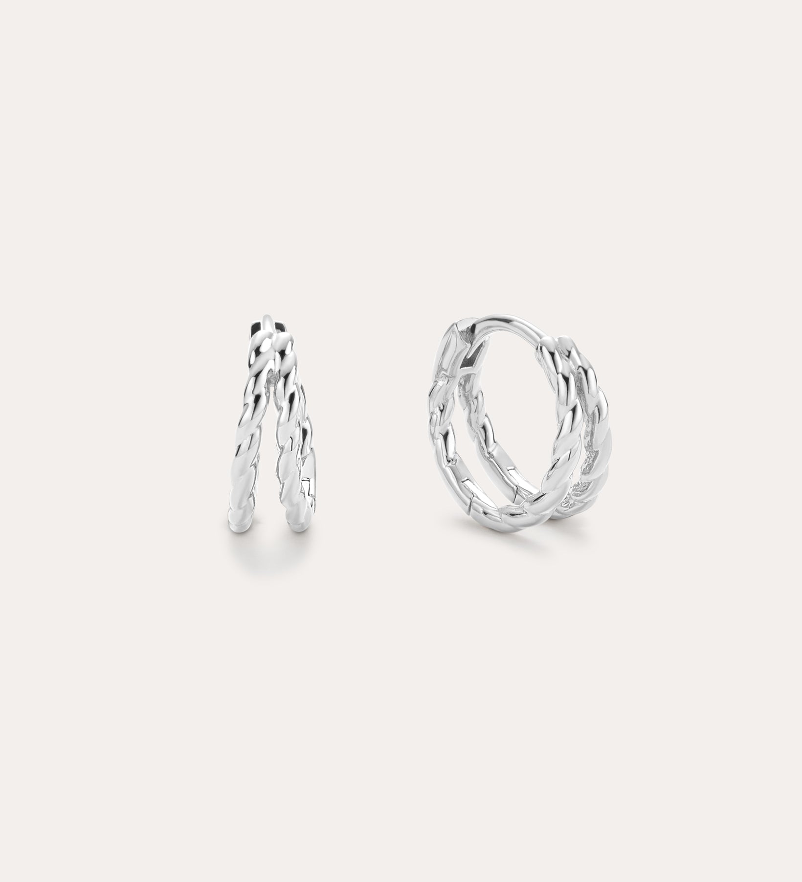 The Mae Twist Double Huggie Earrings in Silver feature a twisted, leafy design. Displayed on a plain background, one earring stands slightly open while the other lies flat, showcasing their intricate detailing.