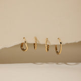 Four 14K recycled solid gold twisted huggie earrings are displayed in a row, hanging through a torn section of beige paper with a neutral background.