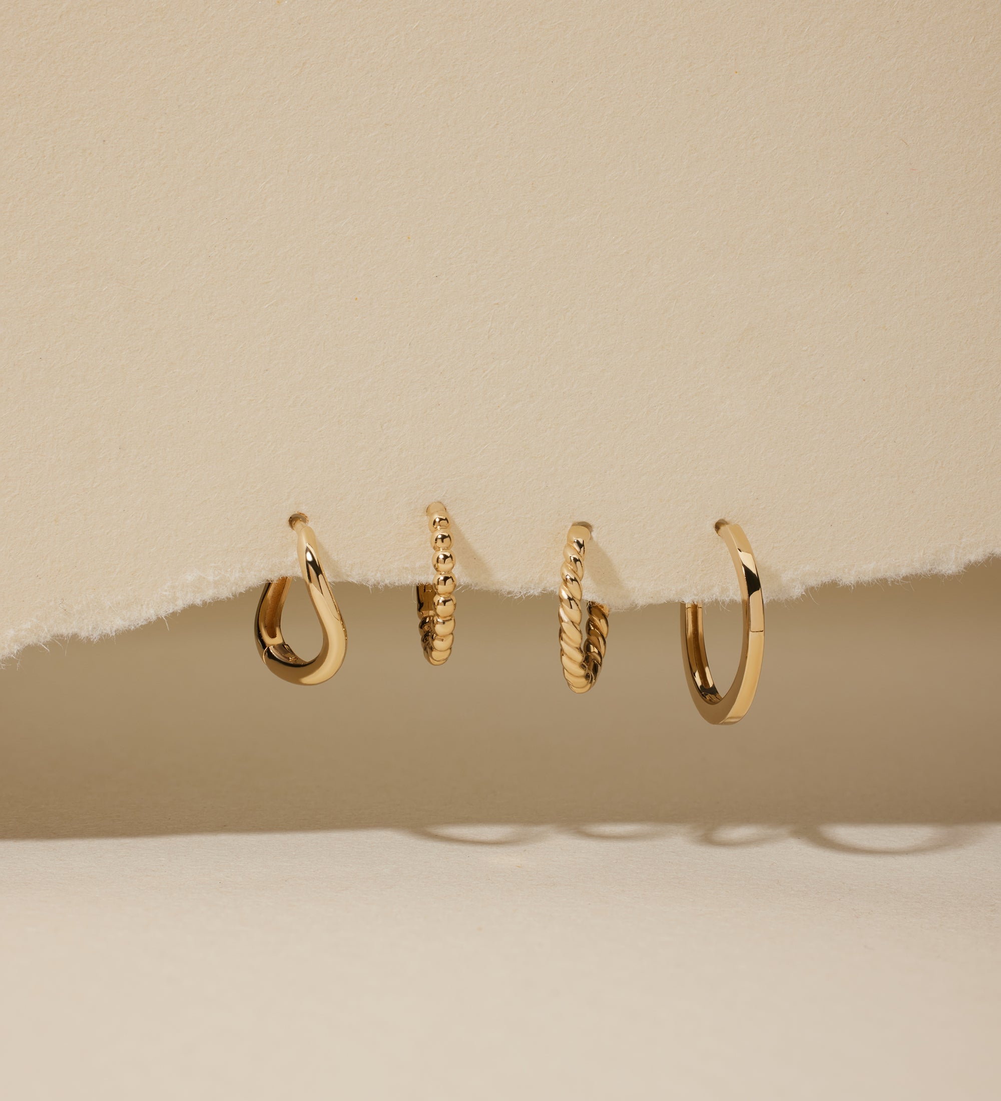 Four 14K recycled solid gold twisted huggie earrings are displayed in a row, hanging through a torn section of beige paper with a neutral background.