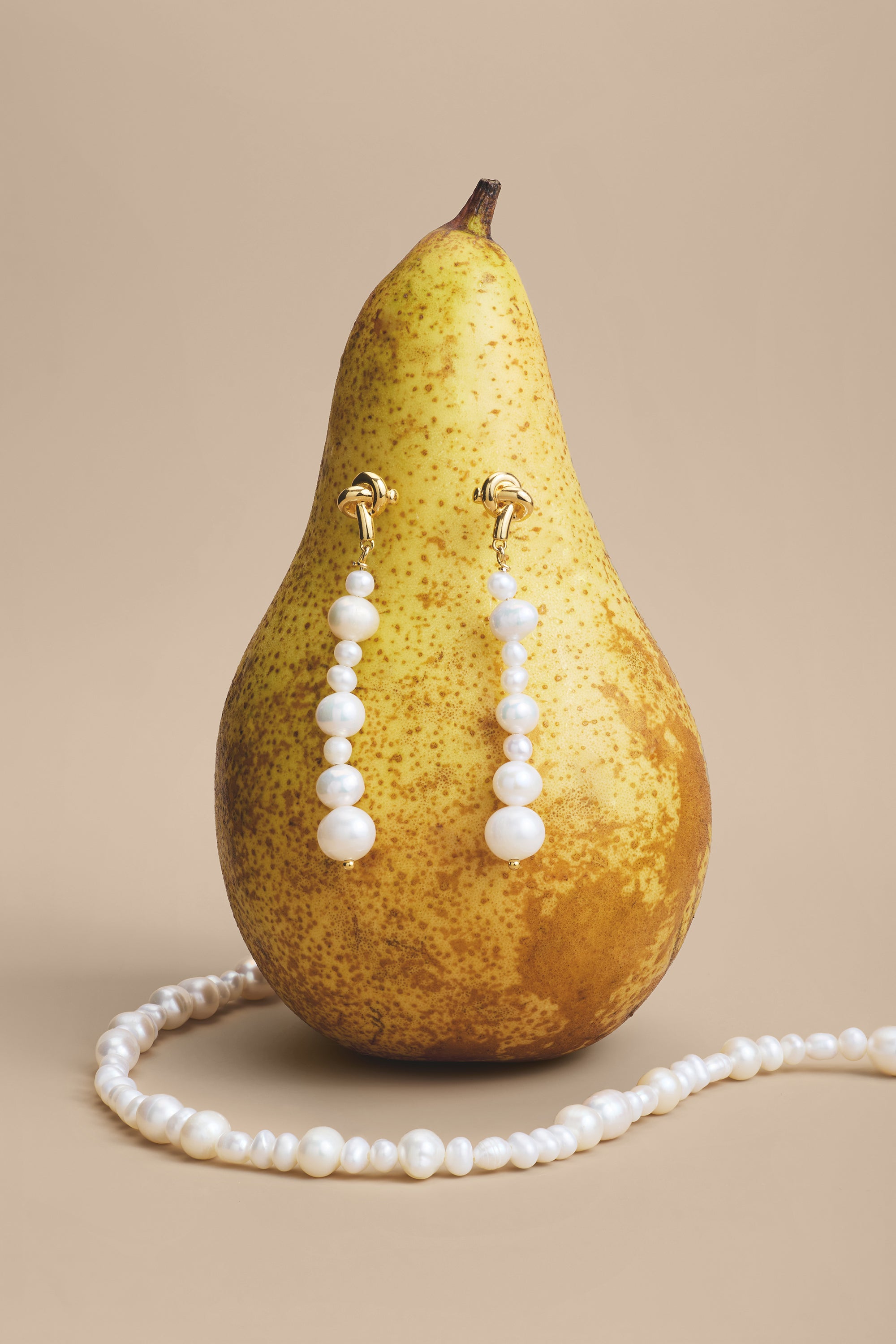 A ripe pear stands upright with pearl drop earrings hanging on it, while the Flora Multi Pearl Toggle Bracelet in Gold—featuring a floral toggle clasp—is coiled at the base. The background is light beige, complementing the colors of the pear and pearls.