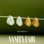A pair of Juno Ridged Chunky Hoops in Silver hangs on a clear rod against a green gradient background. Text below reads, Featured in Vanity Fair.