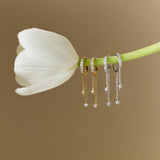 A white tulip with four pairs of earrings—including Star Double Chain Dangle Huggies in Gold—dangling from its stem, set against a plain beige background.