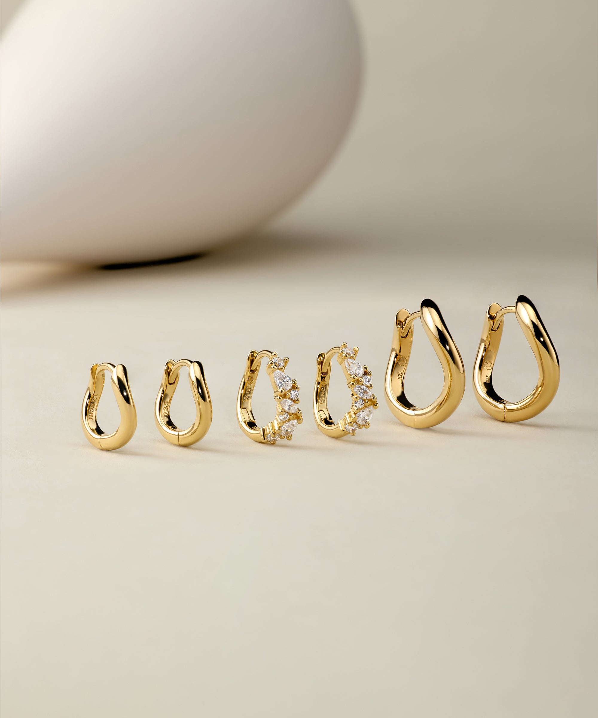 Six pairs of Nori Twisted Hoop Earrings in Gold, including one with embedded crystals, are arranged on a neutral beige surface. A smooth, white, egg-shaped object appears blurred in the background.