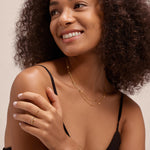 A woman with curly hair in a black tank top smiles to the side, showcasing the Simona Double Chain Stacking Necklace in Gold along with a gold bracelet and rings, set against a plain light background.