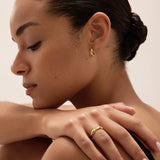 A woman with smooth, dark hair styled in a low bun rests her hand on her shoulder, drawing attention to a silver ring and small gold hoop earring. With natural makeup and pale pink nail polish, she gazes downward with a serene expression—a timeless display of modern jewellery that highlights the elegance of pieces like the Colette Link Droop Ring in Silver.