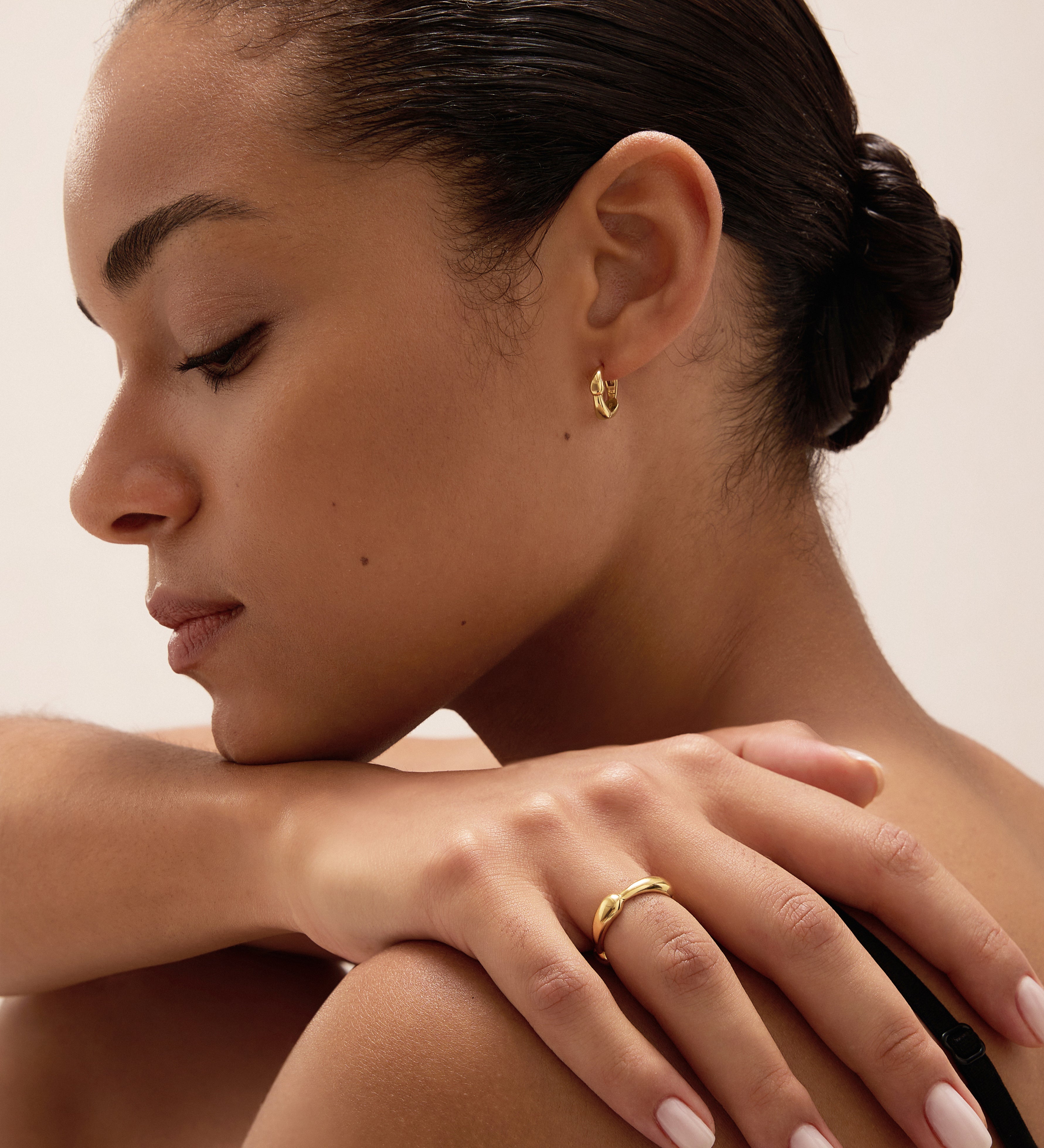 A woman with smooth, dark hair styled in a low bun rests her hand on her shoulder, drawing attention to a silver ring and small gold hoop earring. With natural makeup and pale pink nail polish, she gazes downward with a serene expression—a timeless display of modern jewellery that highlights the elegance of pieces like the Colette Link Droop Ring in Silver.