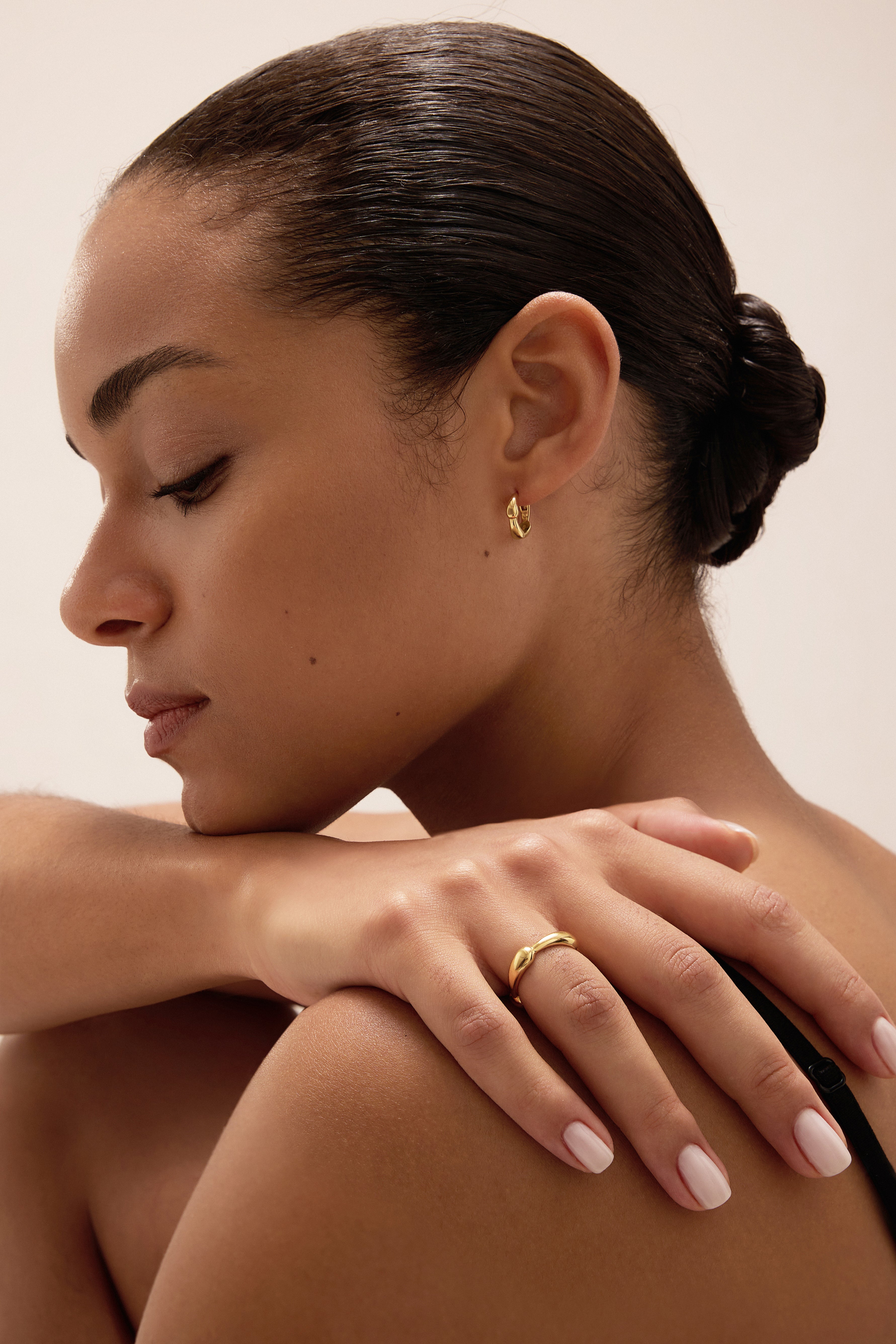 A woman with smooth, dark hair styled in a low bun rests her hand on her shoulder, drawing attention to a silver ring and small gold hoop earring. With natural makeup and pale pink nail polish, she gazes downward with a serene expression—a timeless display of modern jewellery that highlights the elegance of pieces like the Colette Link Droop Ring in Silver.