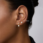 Close-up of a woman’s ear with three earrings: a 2mm 14K solid white gold lab-grown diamond prong set flat back stud in the upper lobe, a medium pearl stud in the middle, and a larger pearl drop earring in the lower lobe. Hair is tied back.