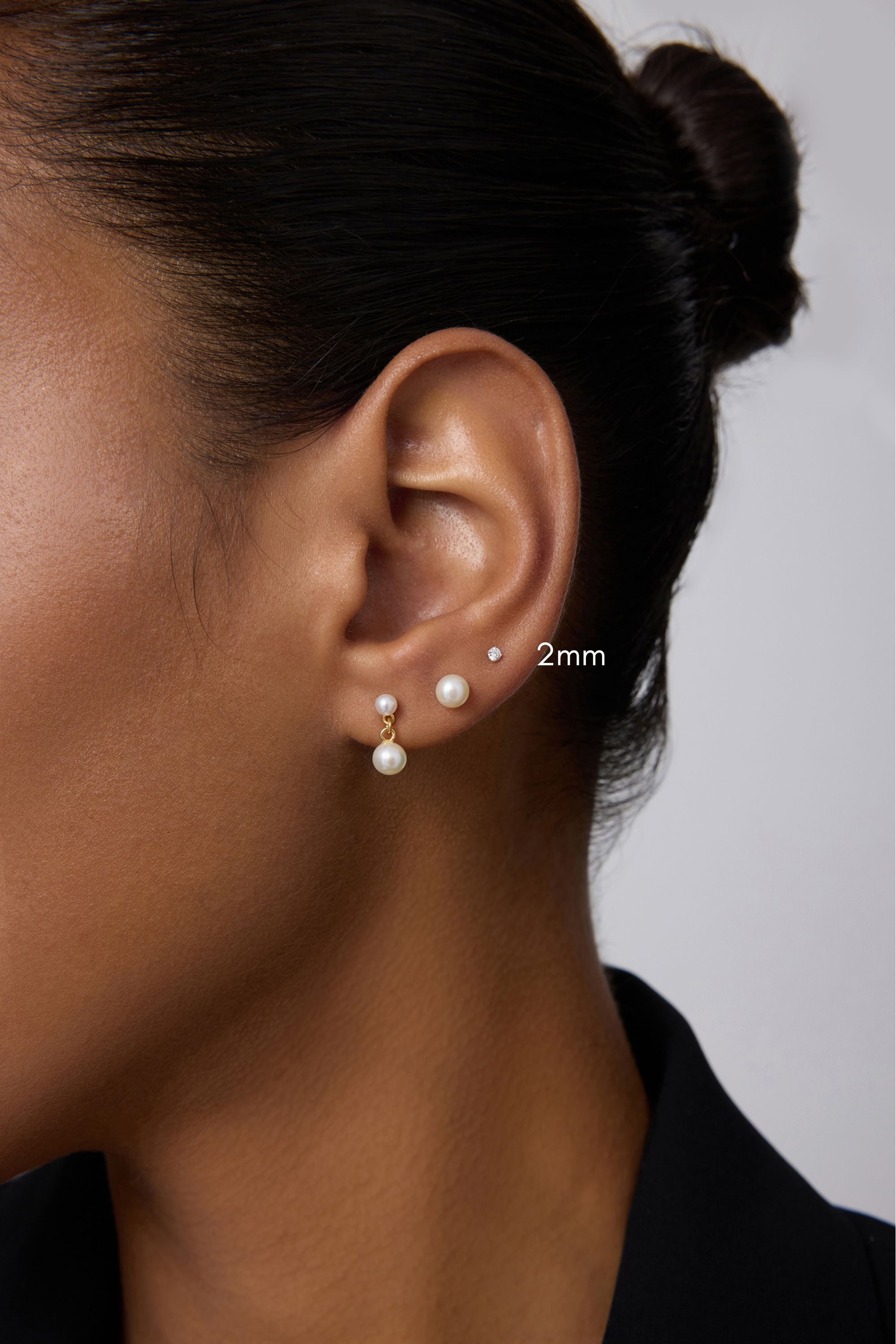 Close-up of a woman’s ear with three earrings: a 2mm 14K solid white gold lab-grown diamond prong set flat back stud in the upper lobe, a medium pearl stud in the middle, and a larger pearl drop earring in the lower lobe. Hair is tied back.