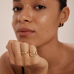 A person with smooth skin rests their chin on their hand, wearing gold hoop earrings and the Colette Link Droop Ring in Silver—a striking example of modern jewellery. They look calmly at the camera against a plain background.
