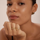 A person with smooth skin rests their chin on their hand, wearing gold hoop earrings and the Colette Link Droop Ring in Silver—a striking example of modern jewellery. They look calmly at the camera against a plain background.
