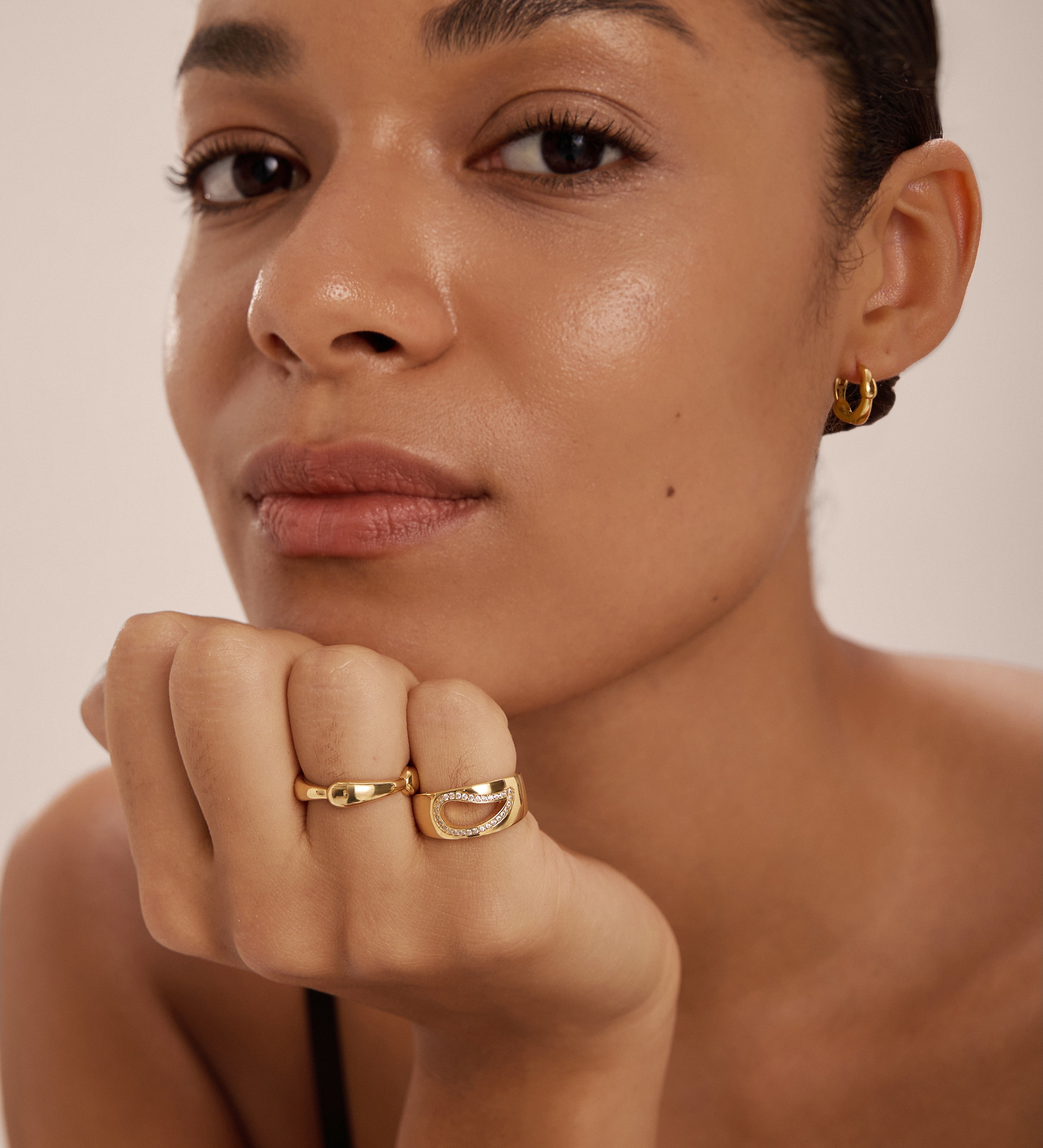 A person with smooth skin rests their chin on their hand, wearing gold hoop earrings and the Colette Link Droop Ring in Silver—a striking example of modern jewellery. They look calmly at the camera against a plain background.