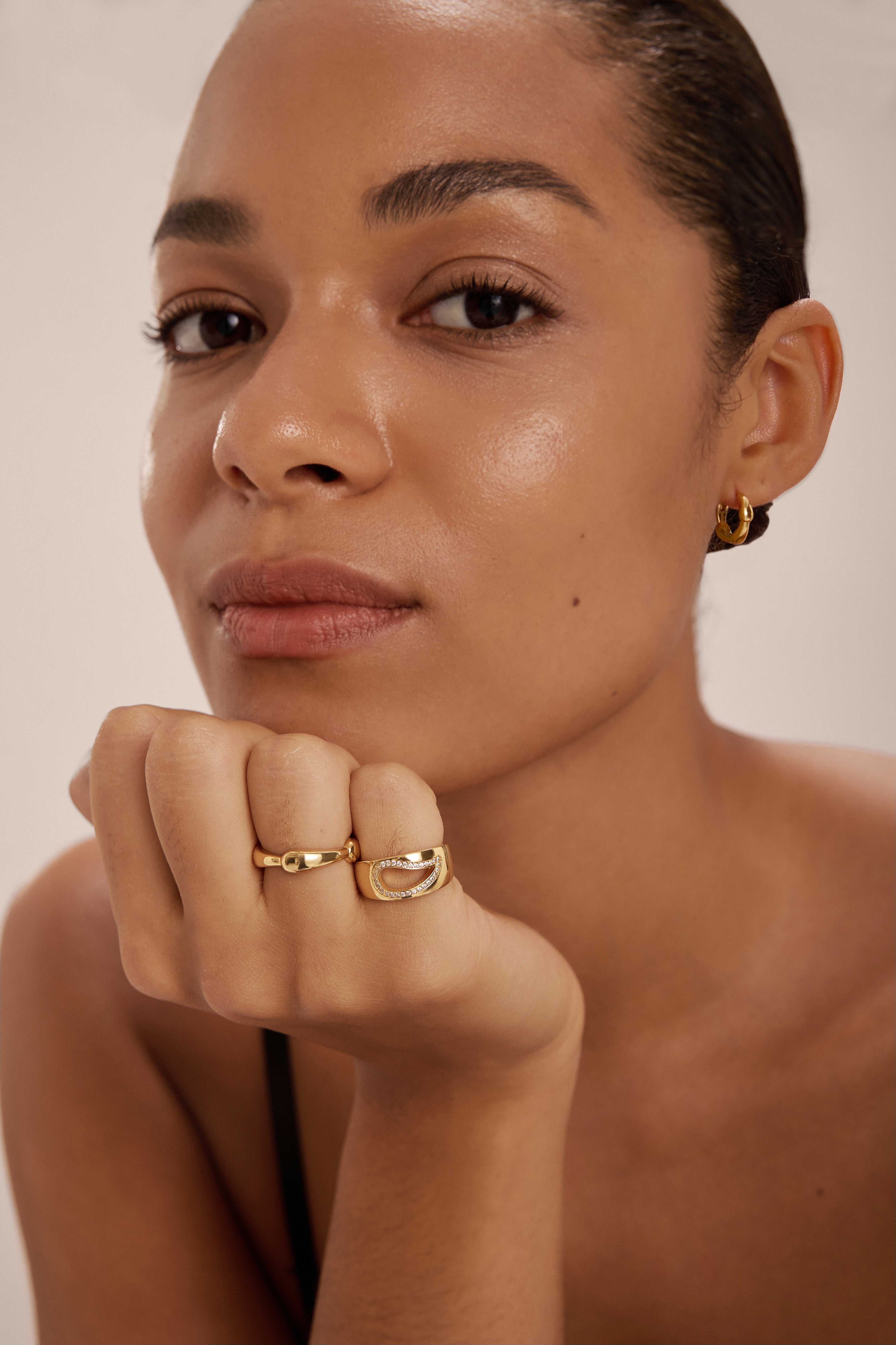 A person with smooth skin rests their chin on their hand, wearing gold hoop earrings and the Colette Link Droop Ring in Silver—a striking example of modern jewellery. They look calmly at the camera against a plain background.