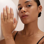 A woman with smooth, medium skin tone wears layered gold jewelry, including the Simona Double Chain Stacking Necklace in Gold. Posing with one hand on a white sphere, she gazes confidently at the camera in a black top and neatly styled hair.