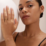 A woman with smooth, medium skin tone wears layered gold jewelry, including the Simona Double Chain Stacking Necklace in Gold. Posing with one hand on a white sphere, she gazes confidently at the camera in a black top and neatly styled hair.