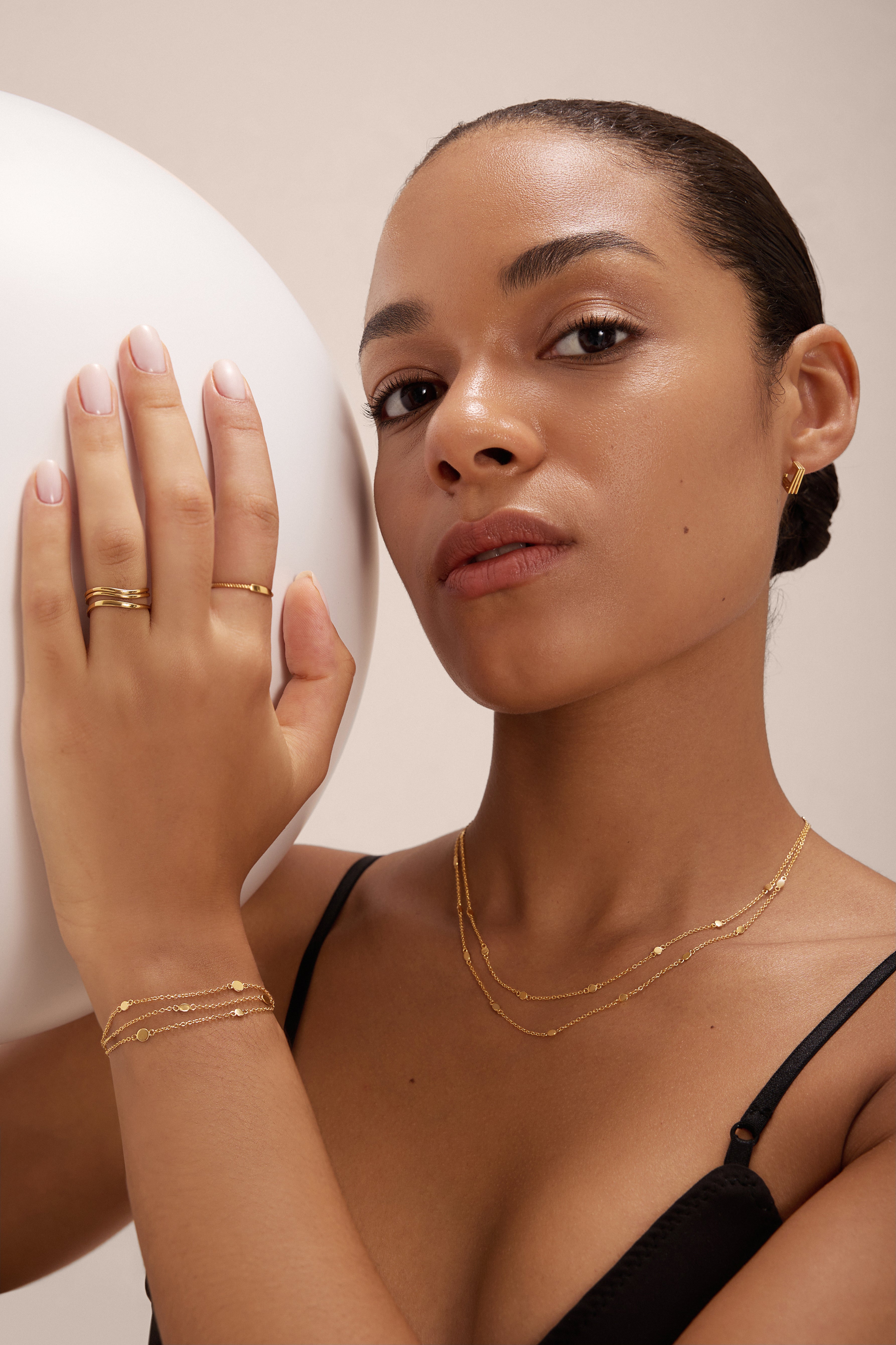 A woman with smooth, medium skin tone wears layered gold jewelry, including the Simona Double Chain Stacking Necklace in Gold. Posing with one hand on a white sphere, she gazes confidently at the camera in a black top and neatly styled hair.