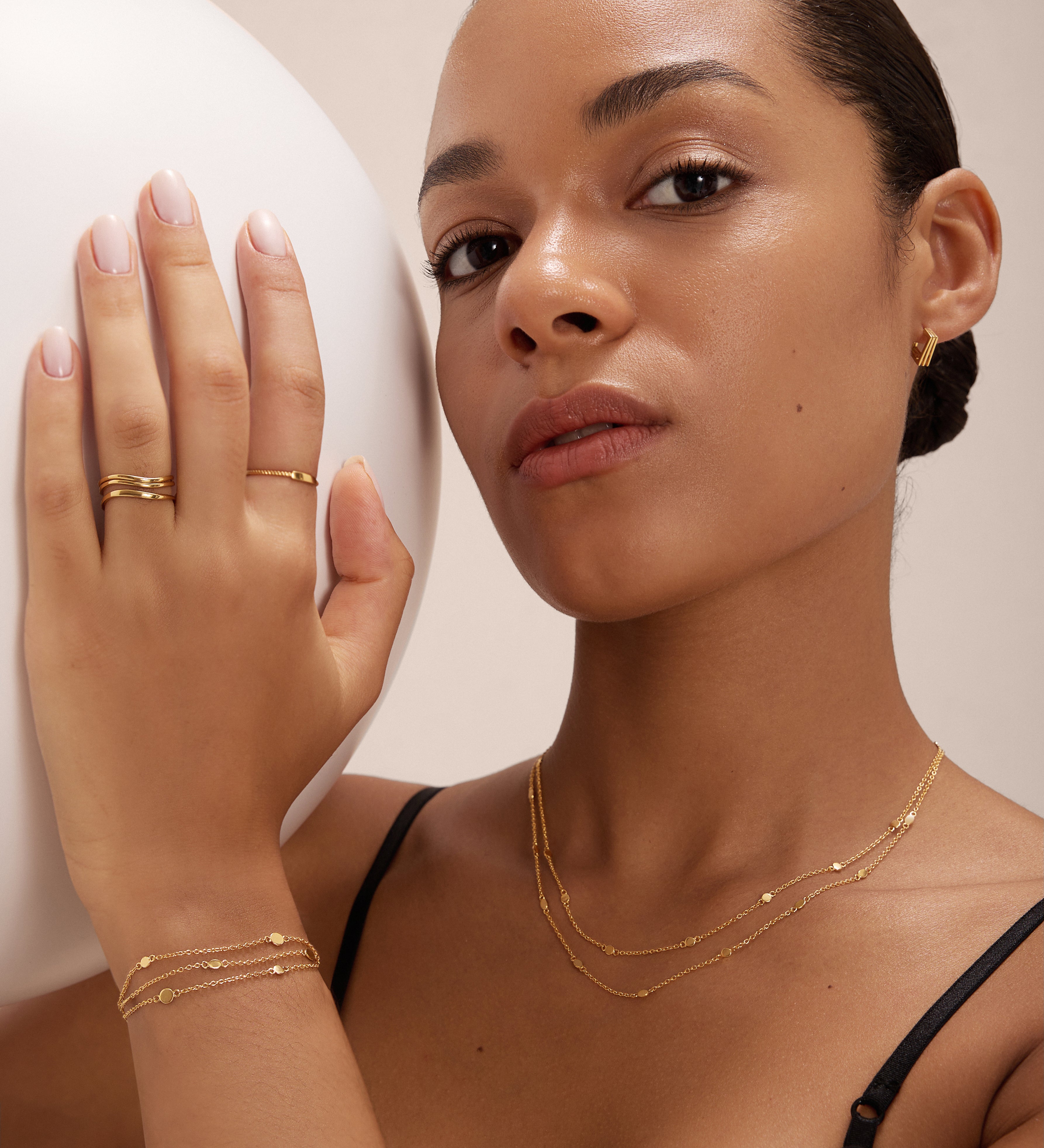 A woman with smooth skin and tied-back hair wears a black top, gold rings, small hoop earrings, and the Simone Triple Chain Layered Bracelet in Gold. She holds a large white sphere in her left hand.