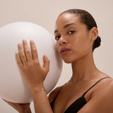 A woman with pulled-back hair, wearing the Simona Double Chain Stacking Necklace in Gold and a black top, confidently holds a large white sphere in one hand while looking at the camera against a neutral background.