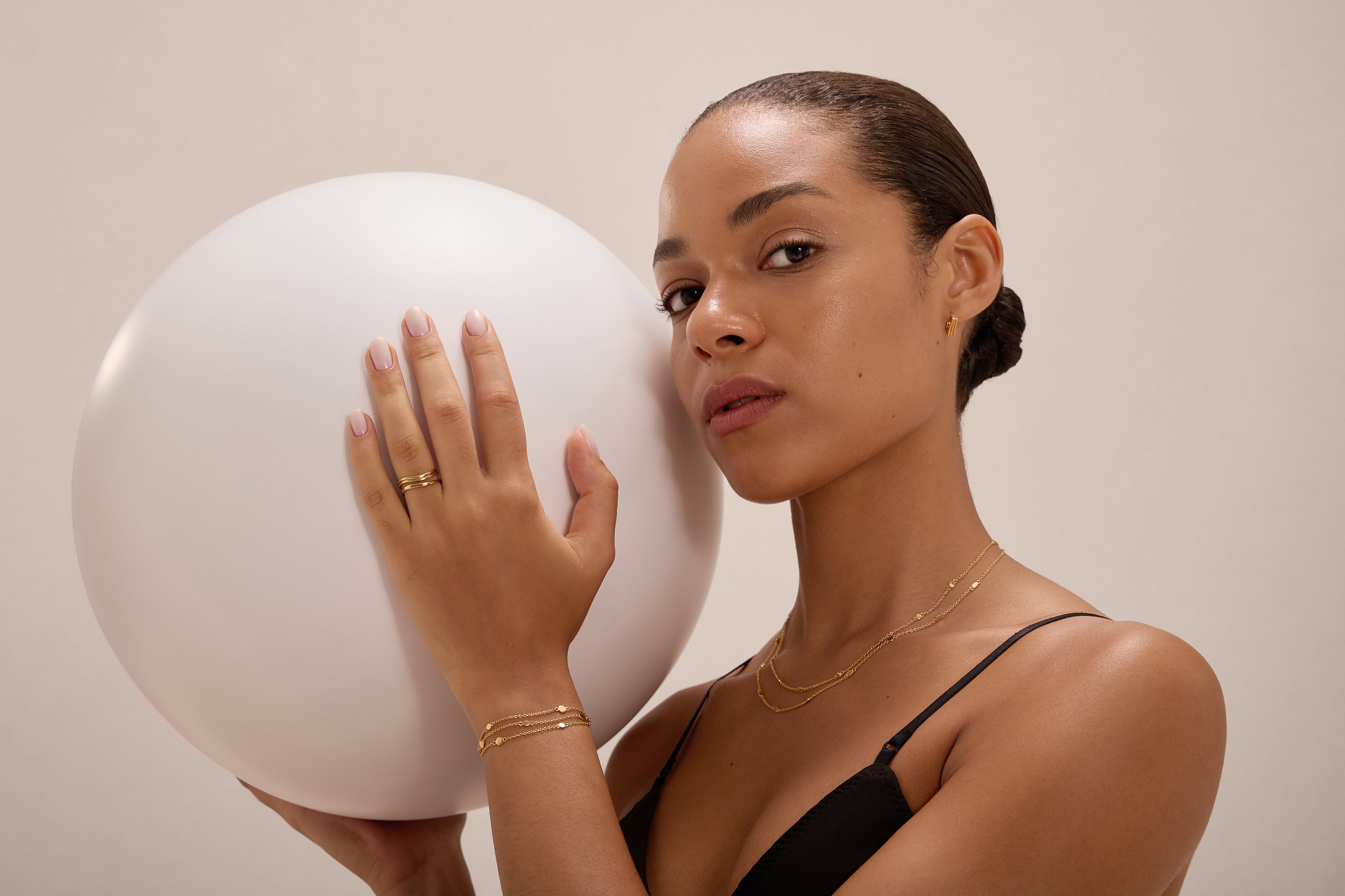 A woman with pulled-back hair, wearing the Simona Double Chain Stacking Necklace in Gold and a black top, confidently holds a large white sphere in one hand while looking at the camera against a neutral background.