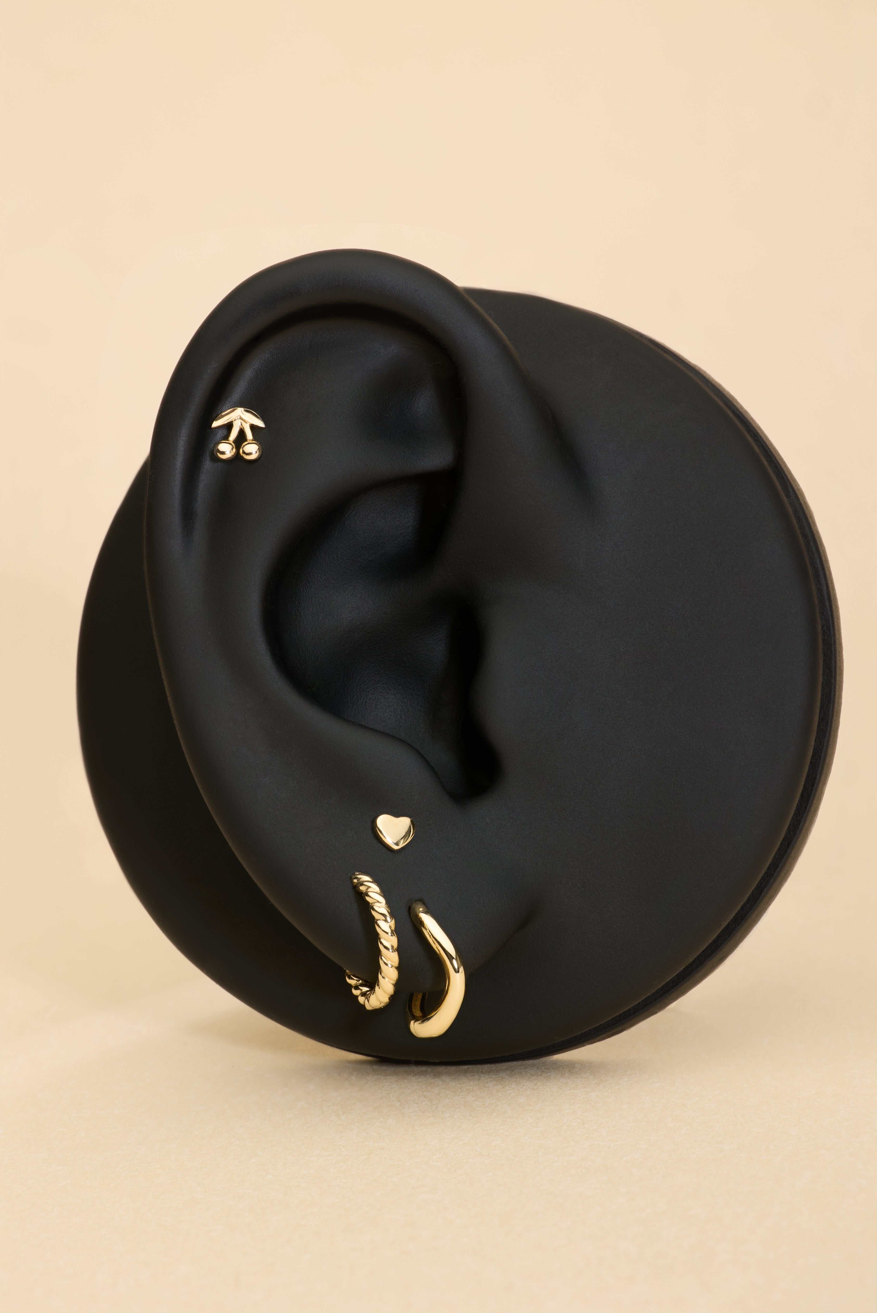 Black ear with gold stud cherry earrings and gold heart flat back stud on a beige background