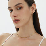 A woman with straight brown hair and natural makeup wears the Estelle Herringbone Chain Necklace in Gold with a white spaghetti strap top, posing against a plain light background.
