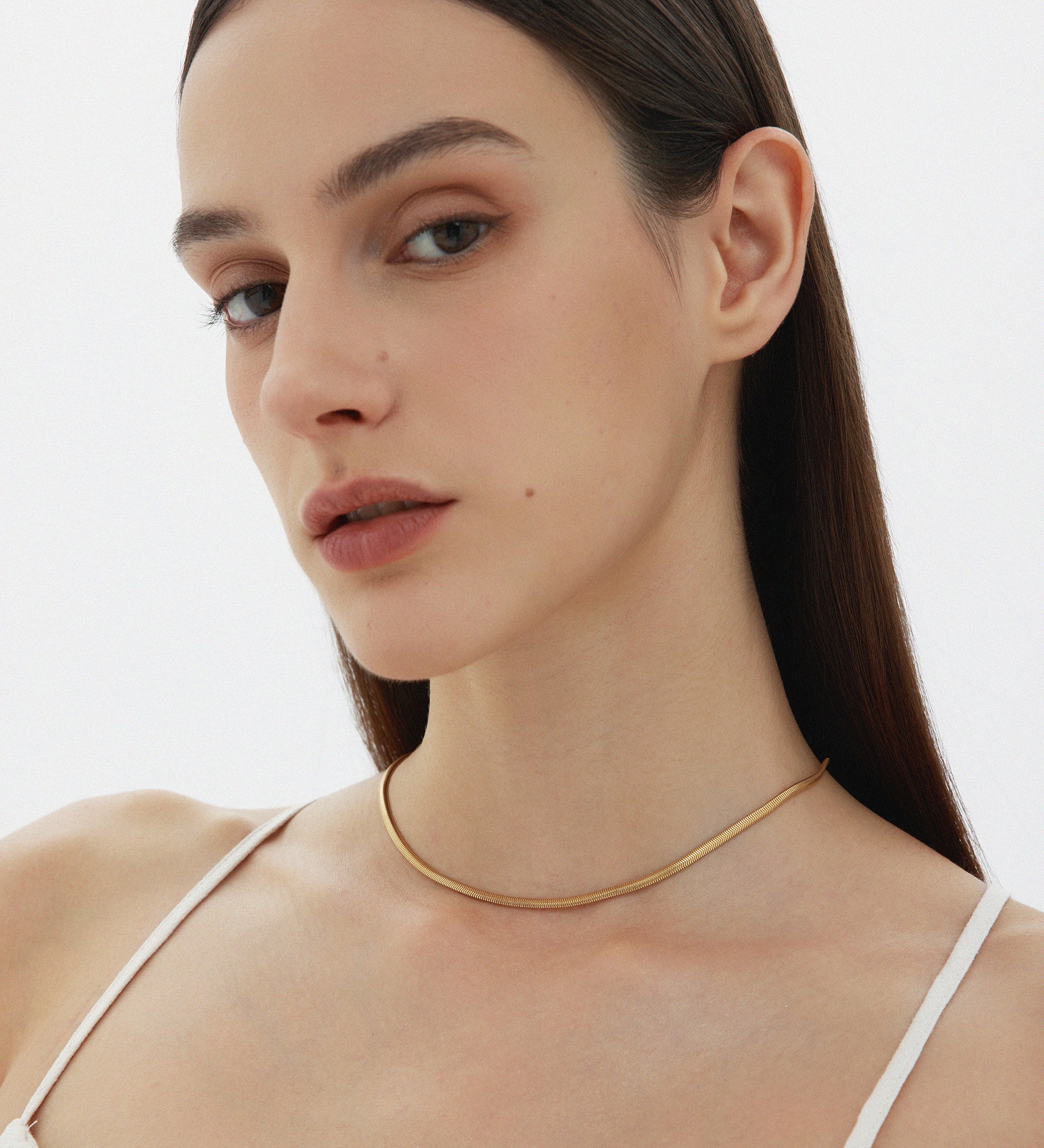 A woman with straight brown hair and natural makeup wears the Estelle Herringbone Chain Necklace in Gold with a white spaghetti strap top, posing against a plain light background.