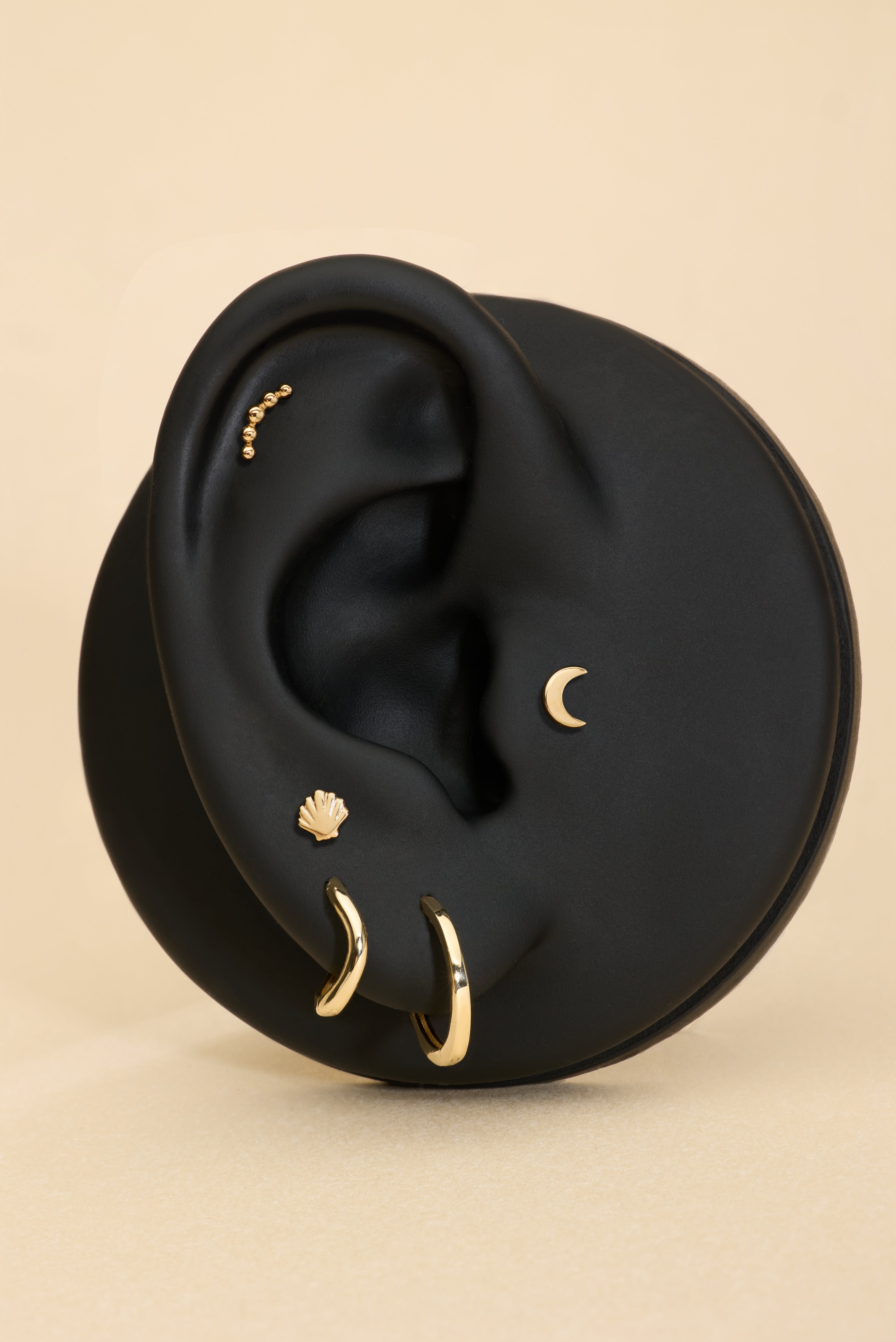 A black ear model showcases the 14K Recycled Solid Yellow Gold Plain Huggie Earring and other sustainable jewelry, set against a beige background.