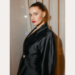 A woman with fair skin and dark hair in a sleek bun wears a shiny black jacket and the Vina Crystal Bezel Ear Cuff in Silver, standing before beige curtains. Text above reads “SPOTTED ON:” and below is the name “GENEVIEVE CHENNEOUR.”.
