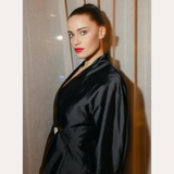 A woman with fair skin and dark hair in a sleek bun wears a shiny black jacket and the Vina Crystal Bezel Ear Cuff in Silver, standing before beige curtains. Text above reads “SPOTTED ON:” and below is the name “GENEVIEVE CHENNEOUR.”.
