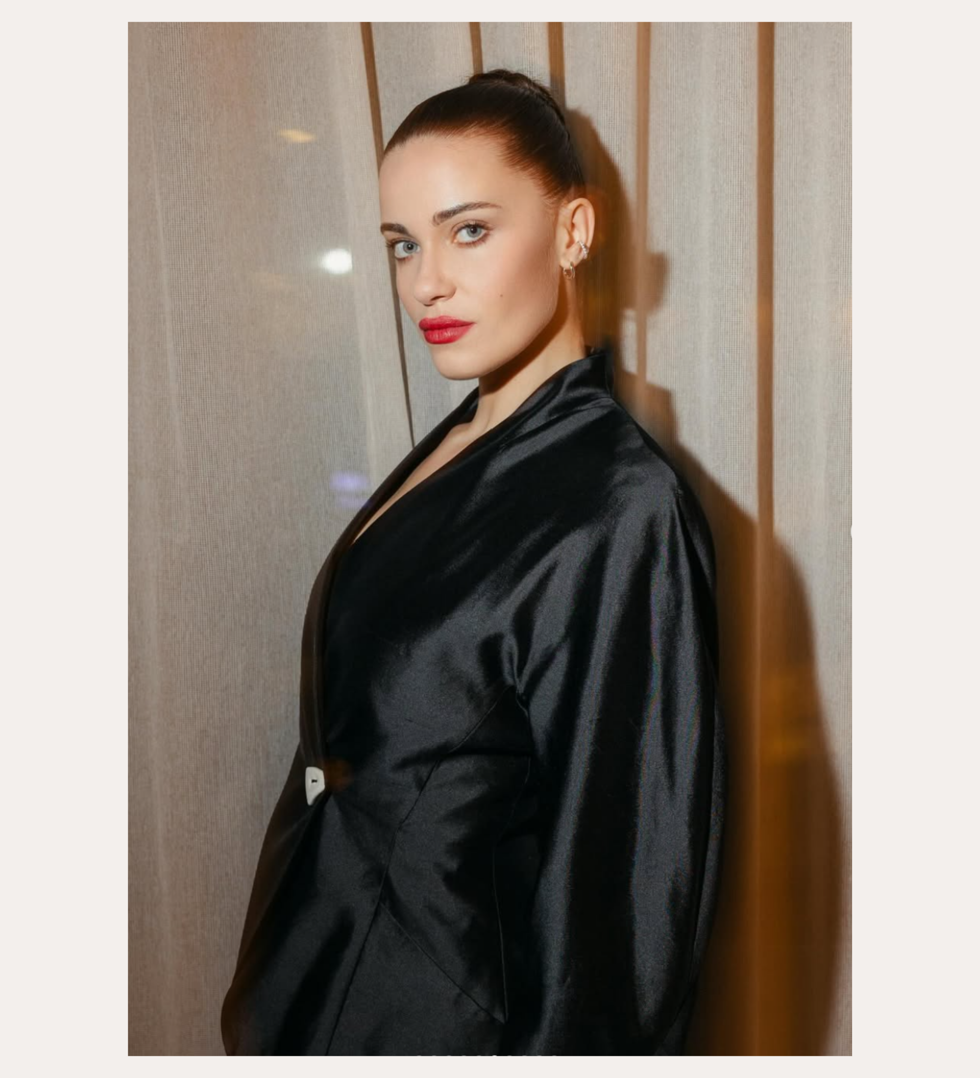 A woman with fair skin and dark hair in a sleek bun wears a shiny black jacket and the Vina Crystal Bezel Ear Cuff in Silver, standing before beige curtains. Text above reads “SPOTTED ON:” and below is the name “GENEVIEVE CHENNEOUR.”.