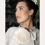 A woman with dark hair in an elegant updo, wearing a textured white outfit with a large flower and bow at the collar, looks to the side. Spotted On: Mimi Webb in Gia Reversible 2 Tone Chubby Hoop Earrings.