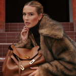 A woman wearing a brown faux fur coat and Georgie Crystal Knife Edge Huggie Hoop Earrings in Gold holds a large brown leather handbag while sitting on steps.