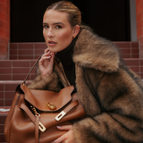 A woman wearing a brown faux fur coat and Georgie Crystal Knife Edge Huggie Hoop Earrings in Gold holds a large brown leather handbag while sitting on steps.