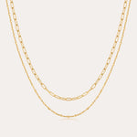 The Mini Paperclip Double Chain Necklace in Gold features two 18k gold plated layered chains—one with delicate elongated links, the other with fine round links—displayed elegantly against a plain, light background.
