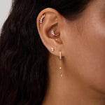 Close-up of a woman's ear with multiple piercings, including gold hoops, cubic zirconia studs, and the Star Double Chain Dangle Huggies in Silver. Her dark wavy hair is partially visible.