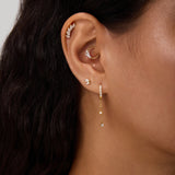 Close-up of a woman's ear with multiple piercings, including gold hoops, cubic zirconia studs, and the Star Double Chain Dangle Huggies in Silver. Her dark wavy hair is partially visible.