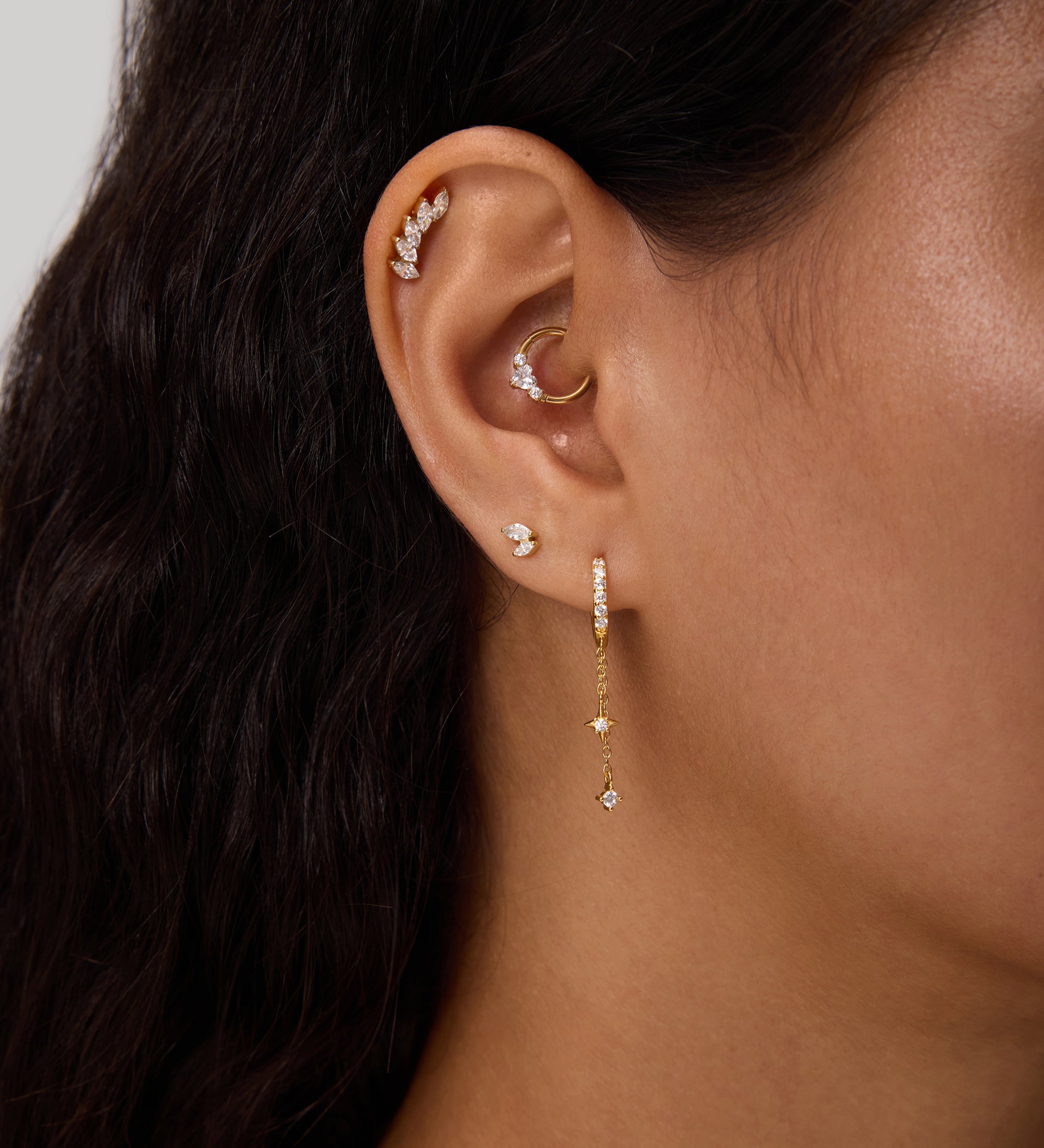 Close-up of a woman's ear with multiple piercings, including gold hoops, cubic zirconia studs, and the Star Double Chain Dangle Huggies in Silver. Her dark wavy hair is partially visible.