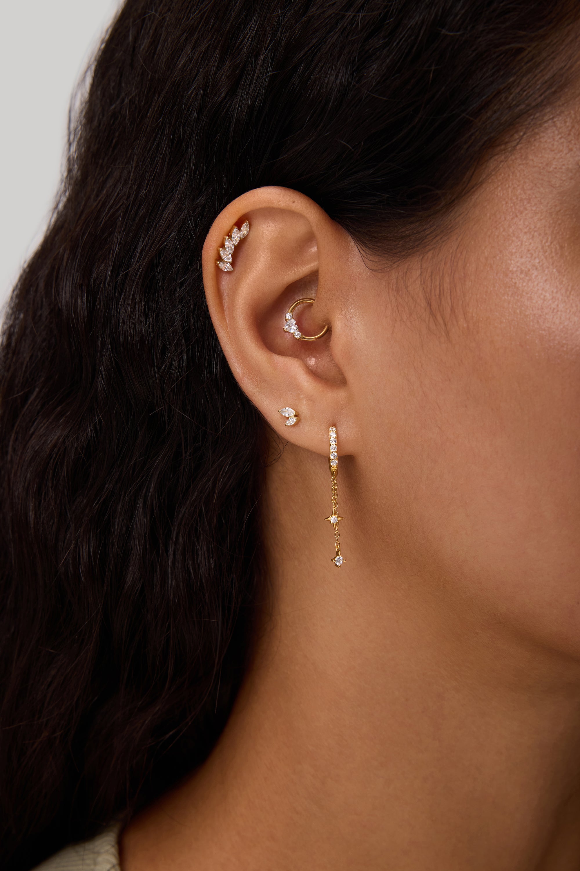 Close-up of a woman's ear with multiple piercings, including gold hoops, cubic zirconia studs, and the Star Double Chain Dangle Huggies in Silver. Her dark wavy hair is partially visible.