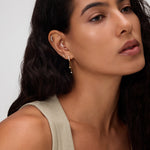 A woman with long, wavy dark hair wears a beige sleeveless top and 18k gold plated Star Double Chain Dangle Huggies in Gold with cubic zirconia. She looks calm, with natural makeup against a neutral background.