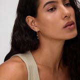 A woman with long, wavy dark hair wears a beige sleeveless top and 18k gold plated Star Double Chain Dangle Huggies in Gold with cubic zirconia. She looks calm, with natural makeup against a neutral background.